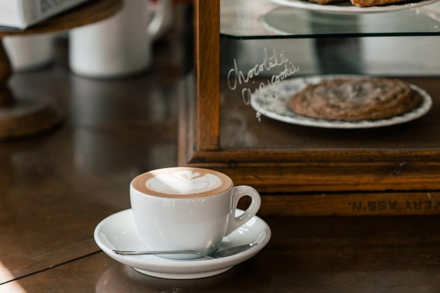 A ceramic mug with a foamy cappuccino beside a silver spoon was placed on a brown wooden table