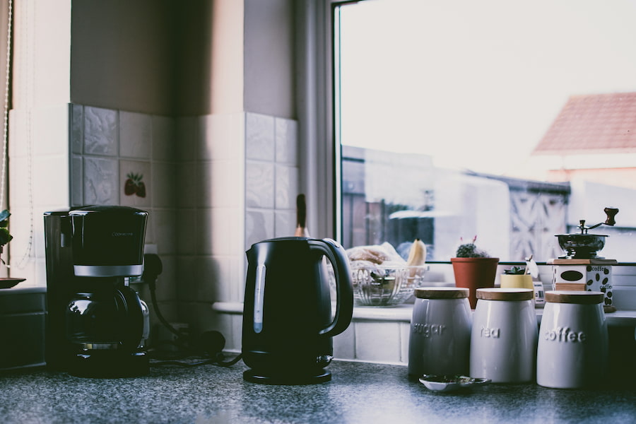 A black coffee maker beside a black water boiler placed on a marbled kitchen top