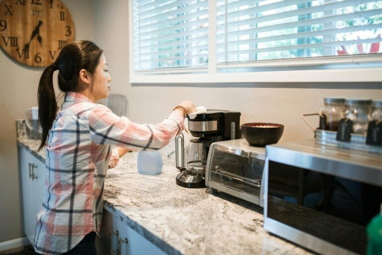 A woman in checkered long sleeves wiping the silver and black coffee maker placed on the kitchen top