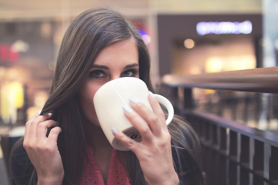 A woman drinking coffee in a white mug seated on a wooden chair at the coffee shop