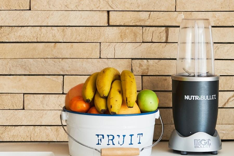 A Nutribullet blender beside different fruits in a white bowl placed on a kitchen top