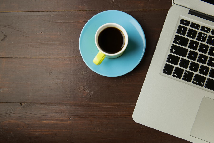 A cup of black coffee in a white mug placed on a blue plate beside a silver laptop on a brown wooden table