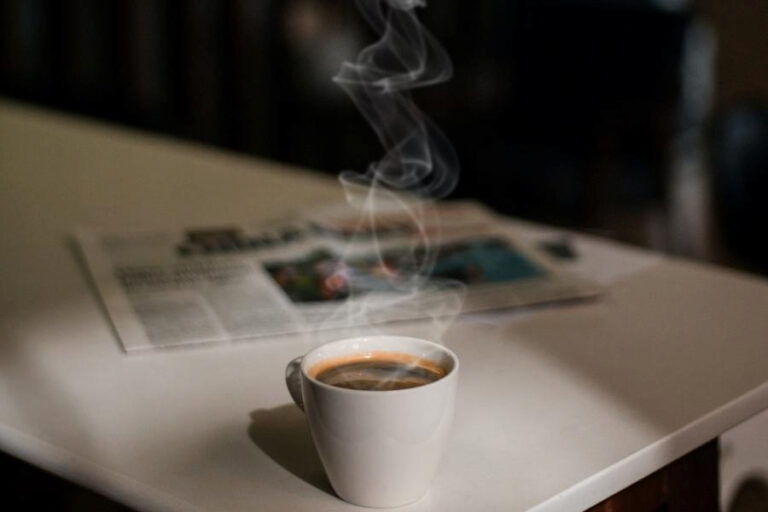 A white cup with hot black coffee beside a newspaper placed on a plastic white table