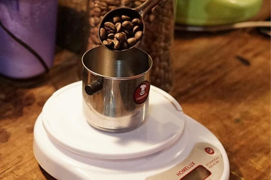 A spoonful of coffee beans being poured on a silver container placed on a white weighing machine