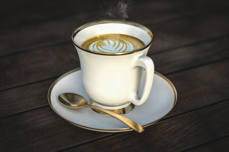 A hot coffee with latte art in a white ceramic cup beside a gold spoon on a white saucer