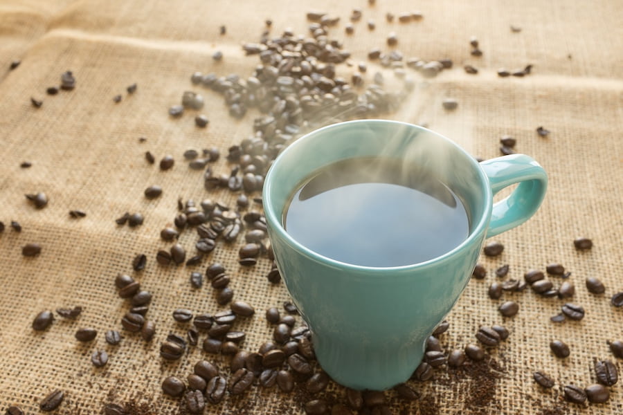 A hot black coffee in a blue cup surrounded by coffee beans is placed on a brown rattan