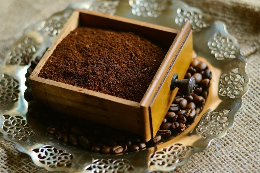 A coffee grounds in a small brown wooden box under coffee beans placed on a round stainless plate