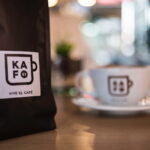 A black pouch bag of KAFO coffee beside a white cup of coffee on a brown wooden table