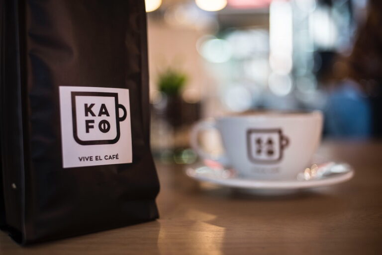 A black pouch bag of KAFO coffee beside a white cup of coffee on a brown wooden table