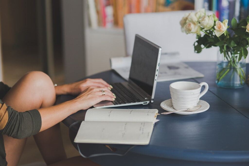 A person using a silver laptop beside a notebook and a white coffee mug placed on a white saucer on a blue table