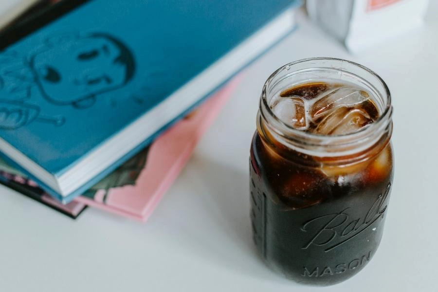 Iced coffee in a mason jar beside a blue and pink notebook was placed on a white surface