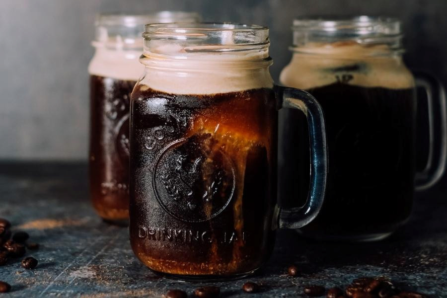 Three mason jars filled with iced cold coffee beside coffee beans on a rough surface