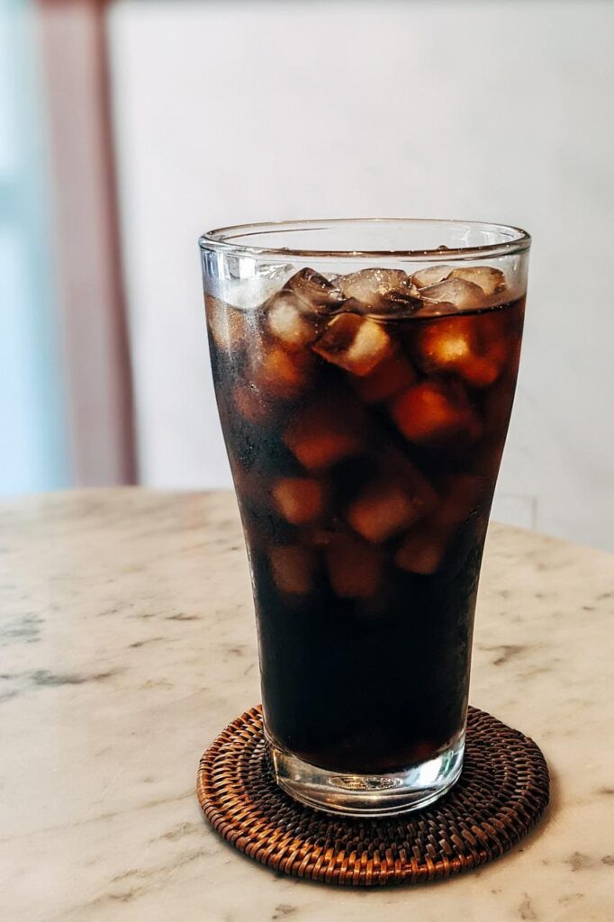 An iced coffee in a glass placed on a brown round-weaved rattan coaster on a white table