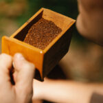 A person holds a small brown wooden box with a coffee ground in it