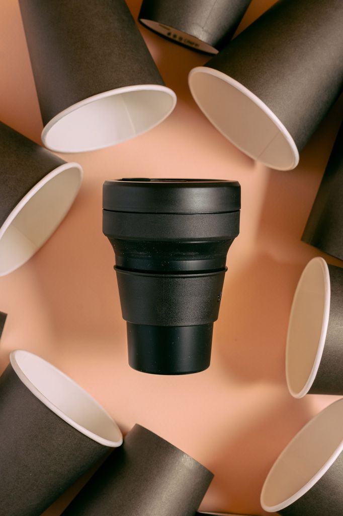 A reusable black cup surrounded by black plastic cups placed on a peach surface