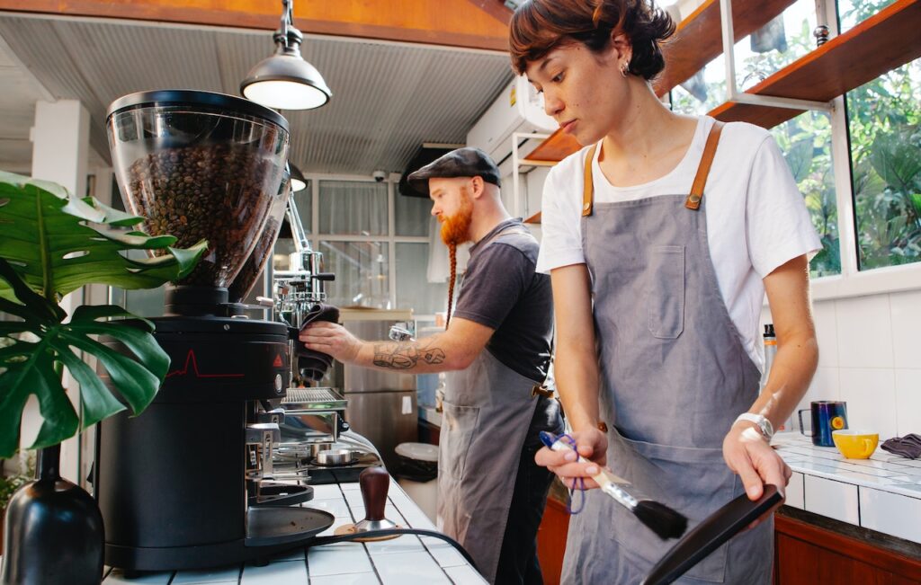 A man holding a black towel wipes the coffee machine while the woman holding a brush cleans the black tray in the coffee shop