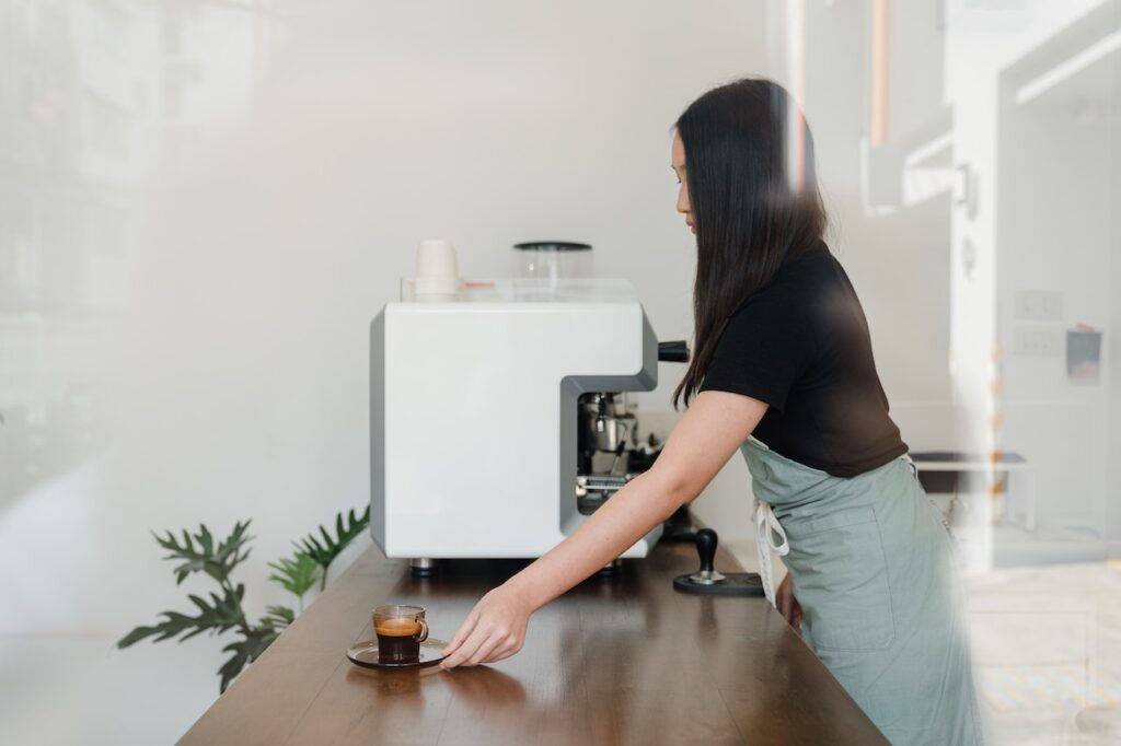 An espresso in a small clear cup is placed on a brown saucer on top of a brown wooden table served by a woman wearing a black shirt and an apron in a coffee shop