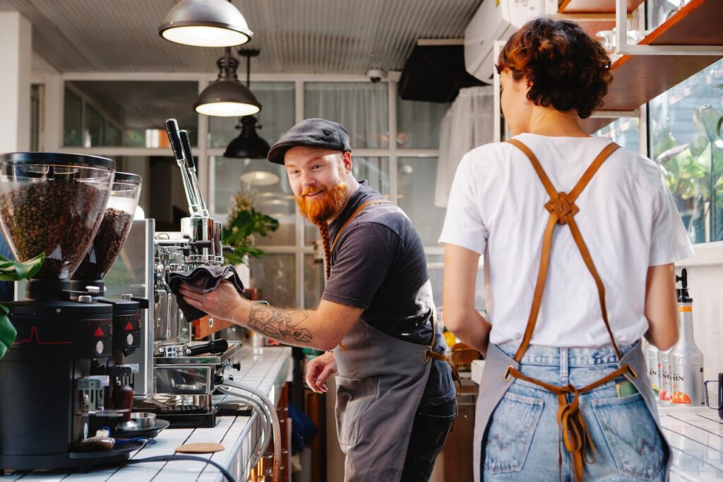 A man holding a black towel wipes the espresso machine while talking to the woman wearing a white shirt in the coffee shop