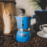 A blue Moka pot beside coffee beans and a white cup of coffee on a white saucer placed on top of a black table