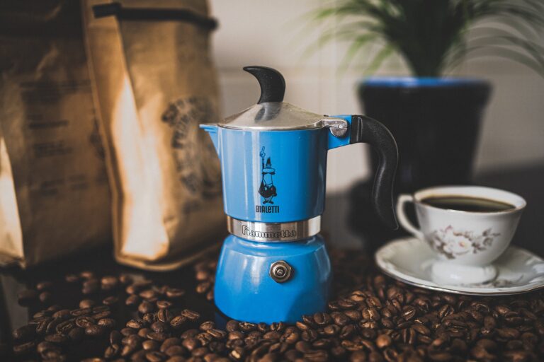 A blue Moka pot beside coffee beans and a white cup of coffee on a white saucer placed on top of a black table