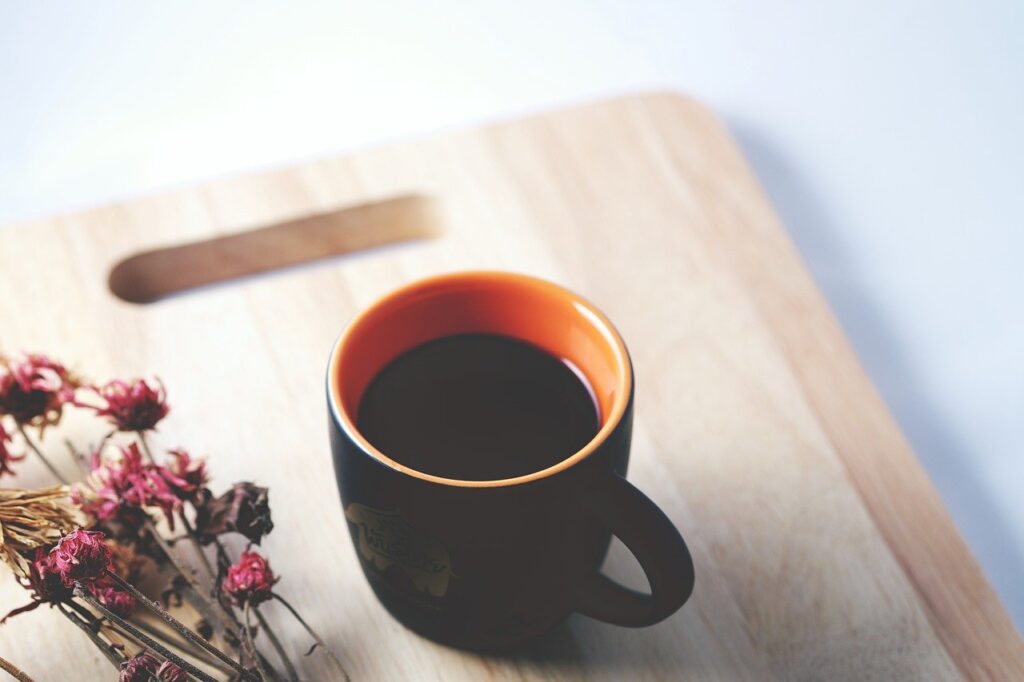 A black cup filled with a coffee beside a dry flower on a brown wooden chopping board