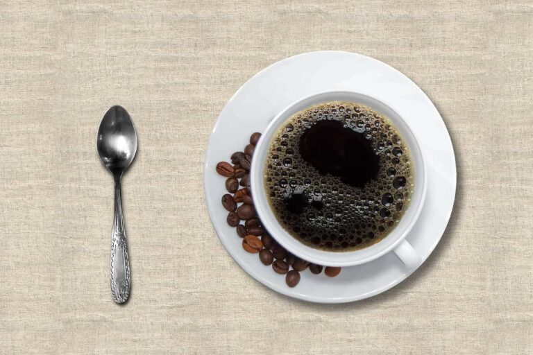 A white cup filled with black coffee was placed on a white saucer beside a silver spoon on a placemat