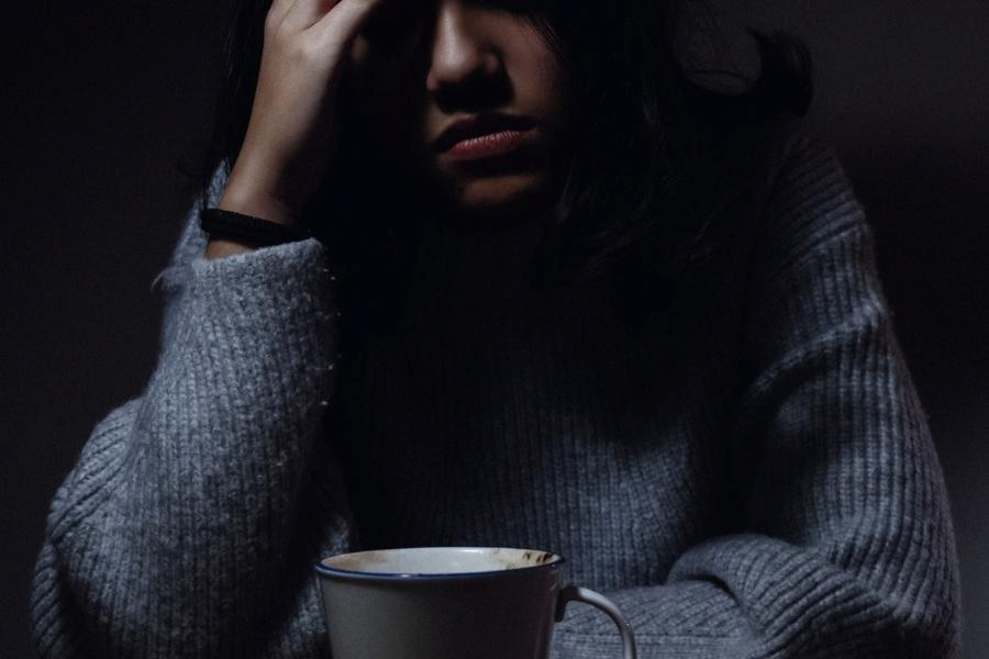 A woman in a blue top holds her head while having a cup of coffee placed in a white mug