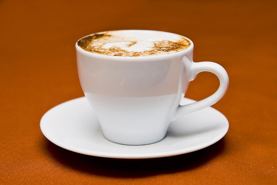 A close-up white cup filled with foamy coffee is placed on a white saucer on an orange surface