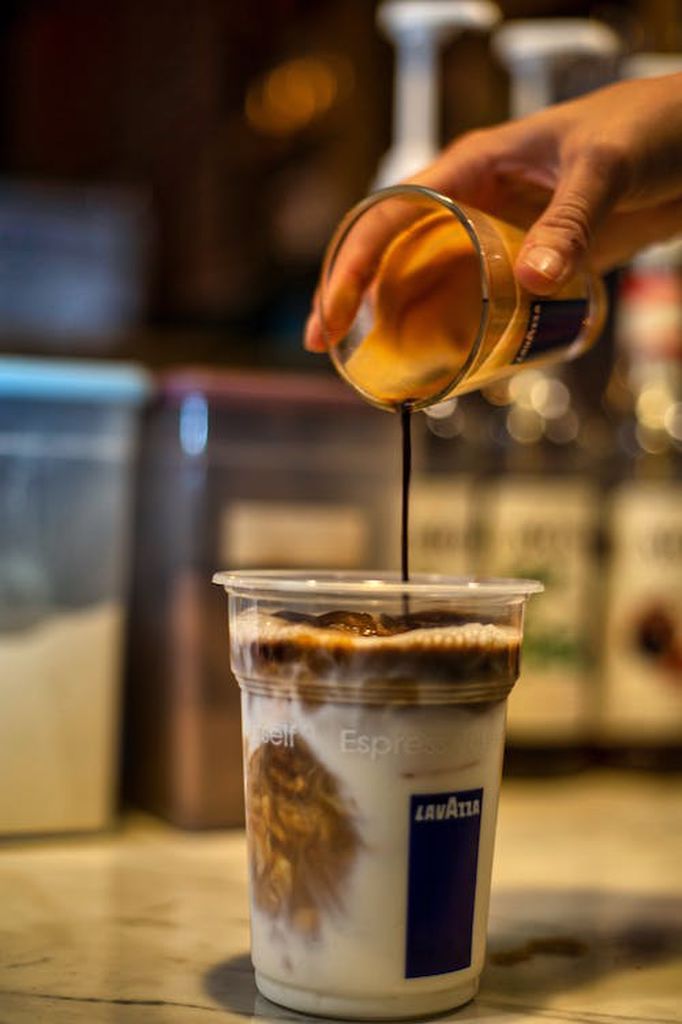 A person pouring syrup on a plastic cup filled with iced coffee placed on a white surface in a coffee shop