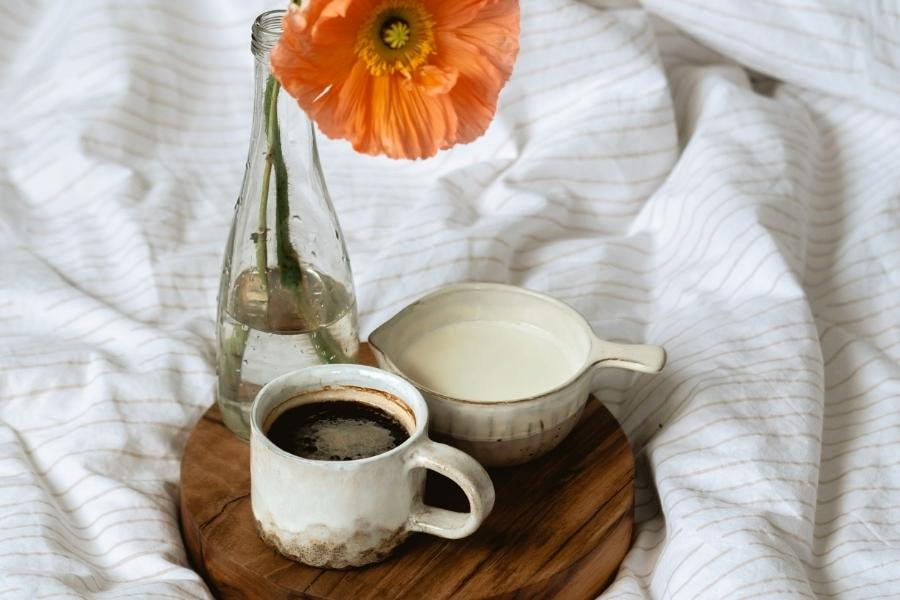 A white cup filled with black coffee beside a cup filled with milk and a clear flower vase on a brown wooden chopping board