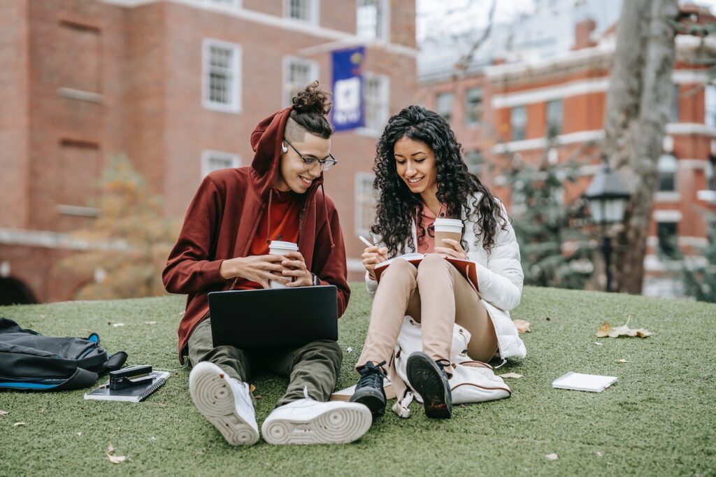 A man and a woman hold cups of coffee while using a laptop and notebook to study seated on green grass