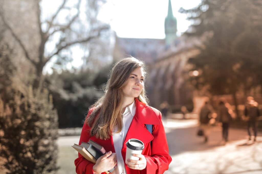 A young woman holds a cup of coffee and three books wearing a white blouse and red coat while walking outside