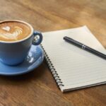 A cup of cappuccino in a blue mug placed on a blue saucer with a spiral notebook and a black ballpen on a brown wooden table