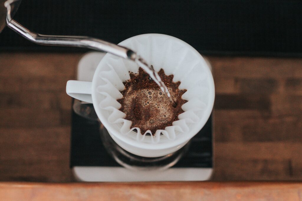 Hot water from a metal container is being poured into the white coffee filter on a brown wooden table