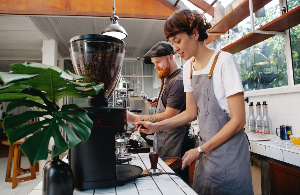 A woman holds a brush cleaning the white tiles while a man fixes the espresso machine in the coffee shop