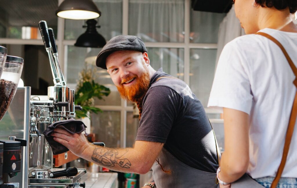 A smiling man wipes the espresso machine using a purple rug while a woman talks to him in a coffee shop