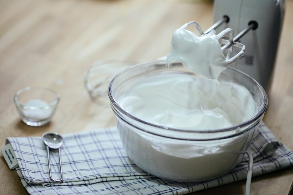 A white whipping cream inside a clear glass bowl on top of a checkered table cloth near a mixer on top of a wooden table