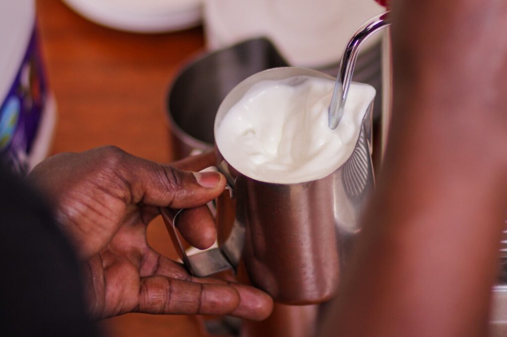 A person holding a stainless container with milk while using the metal frother on top of a brown wooden table