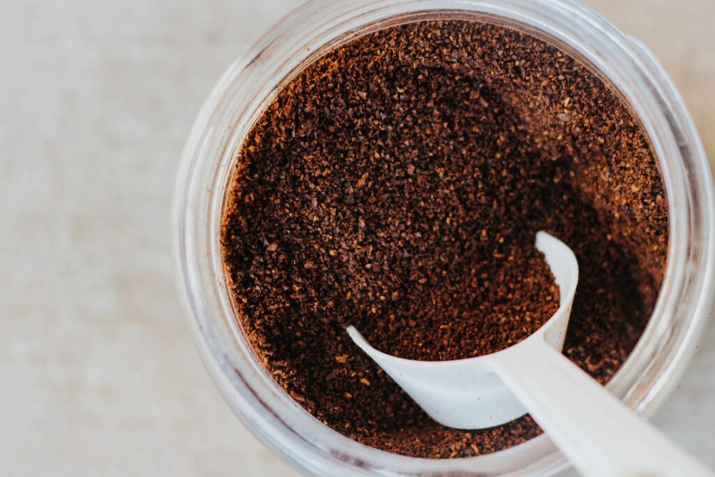 Coffee grounds on a clear container with a white scoop placed on top of a white surface