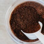 Coffee grounds on a clear container with a white scoop placed on top of a white surface