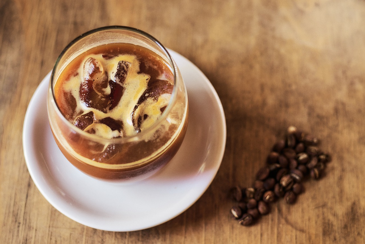 Ice cold coffee in a glass sitting on top of a white plate with coffee beans on the side on the wooden table