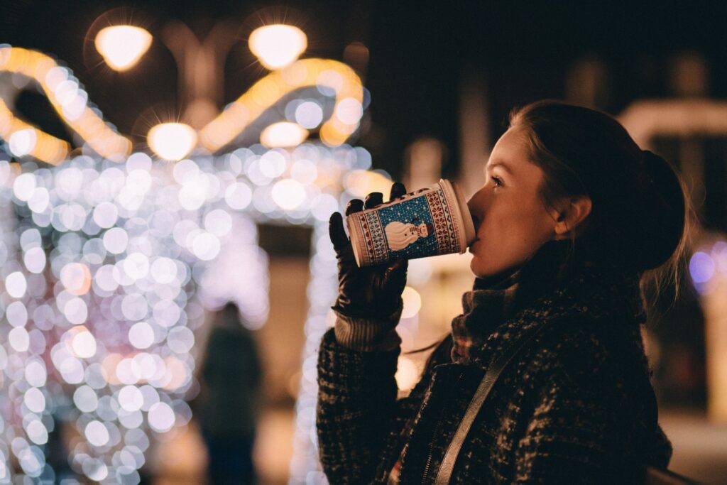 A girl in a ponytail wearing a checkered scarf and black leather gloves is drinking a cup of coffee outside