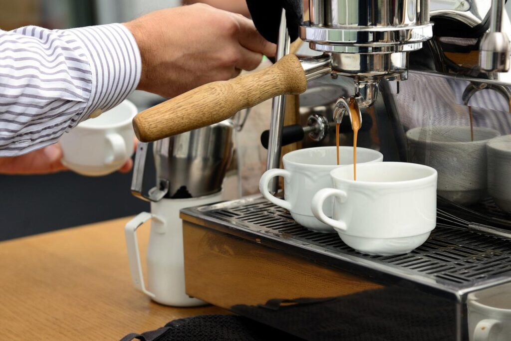 Two fresh cups of espresso being made in a coffee maker by a barista wearing a striped long sleeved shirt in a coffee shop