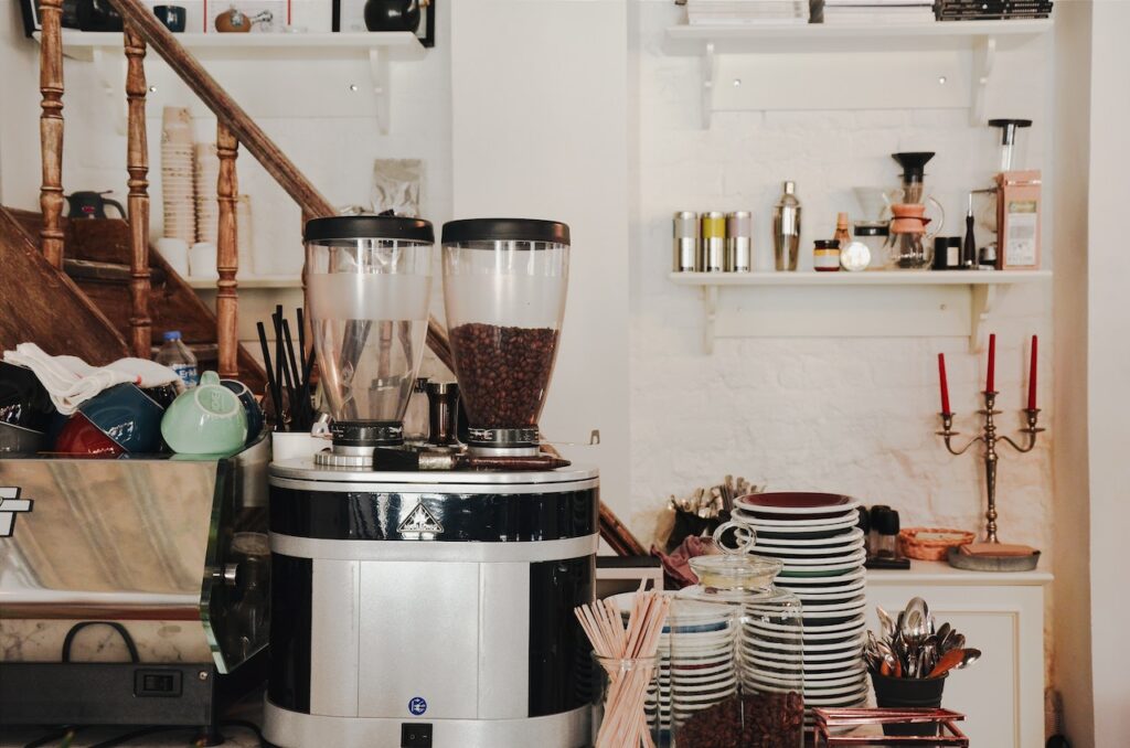 A black and silver coffee grinder near a bundle of wooden stirrers and white saucers on a table near the brown wooden stairs