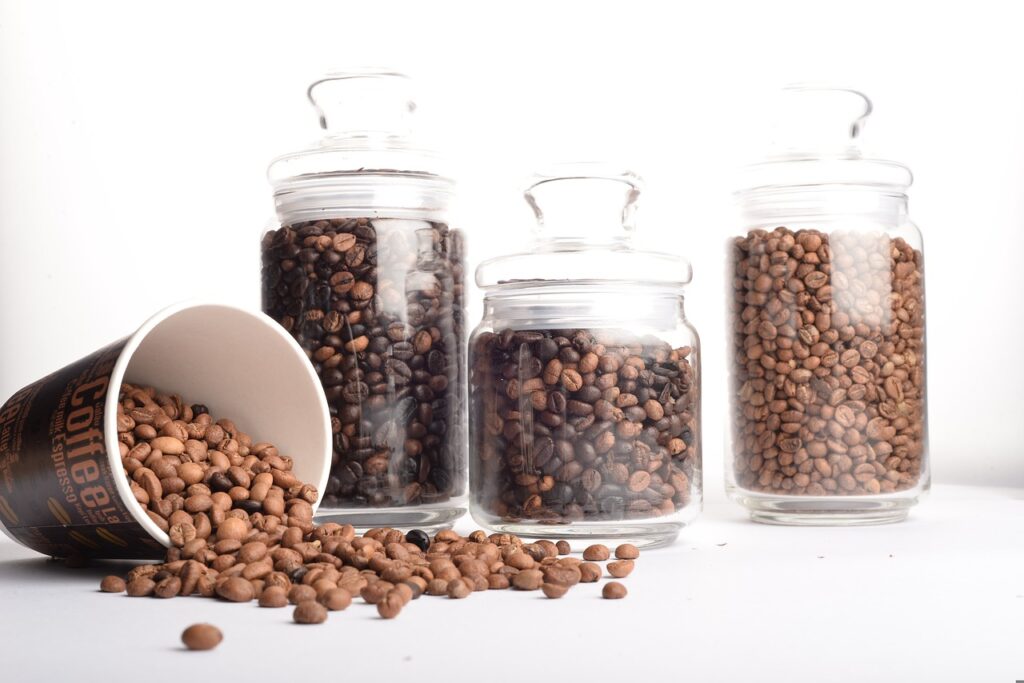 Different coffee beans in three clear glass jars and a coffee cup on a white surface