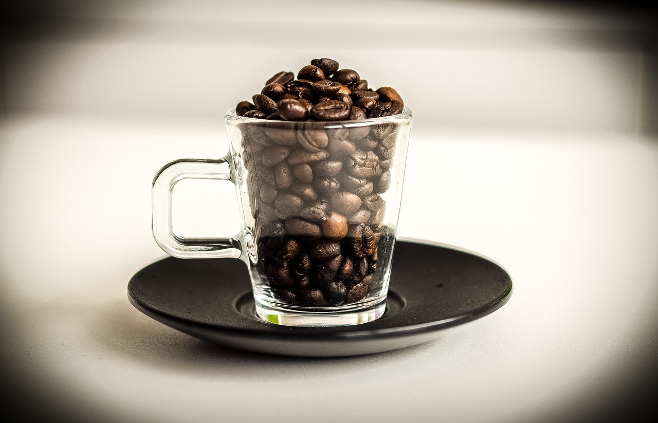 Coffee beans inside a clear coffee mug are placed on a black saucer on a white surface