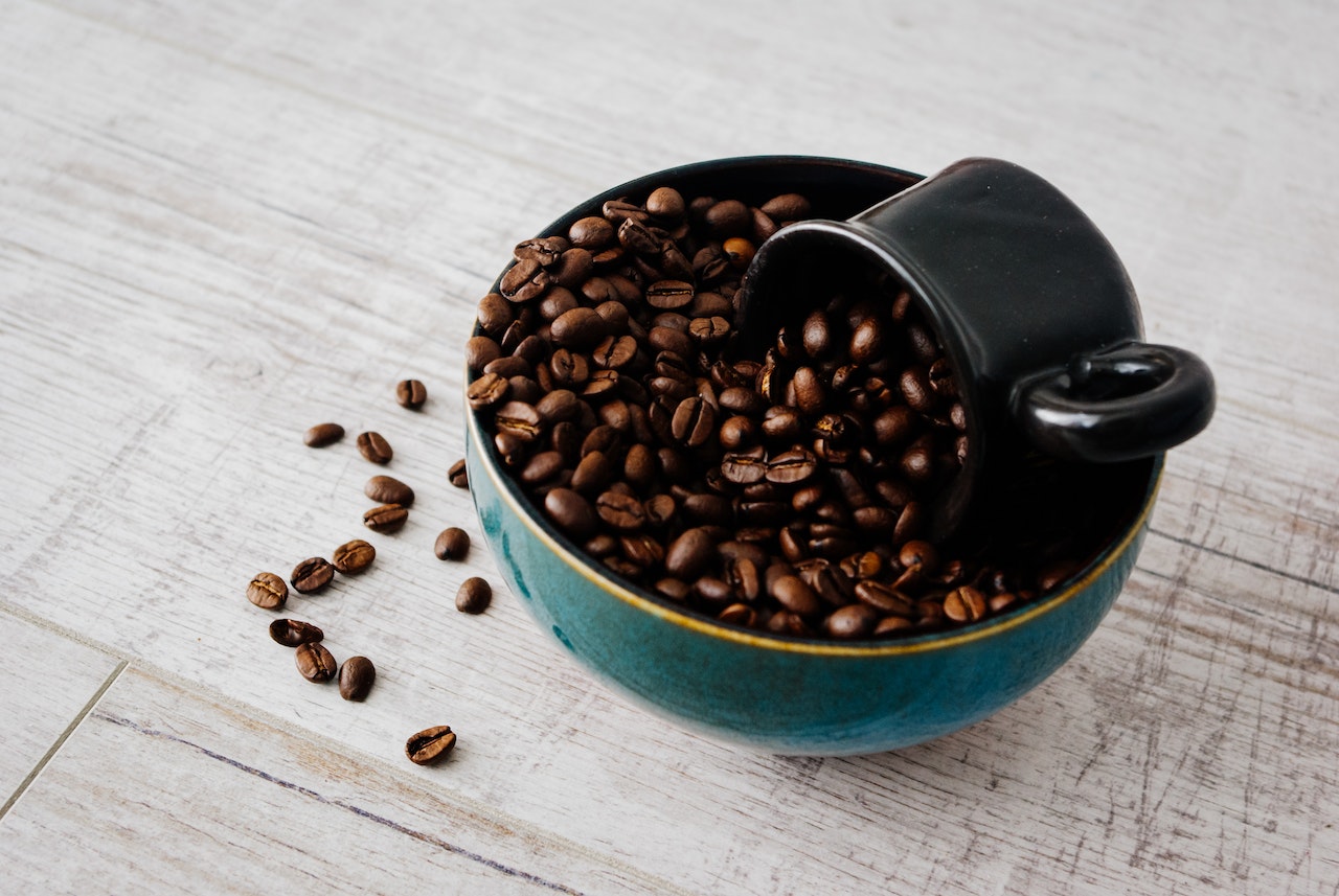 A black mug scooping whole coffee beans placed in a blue bowl on a white wooden table