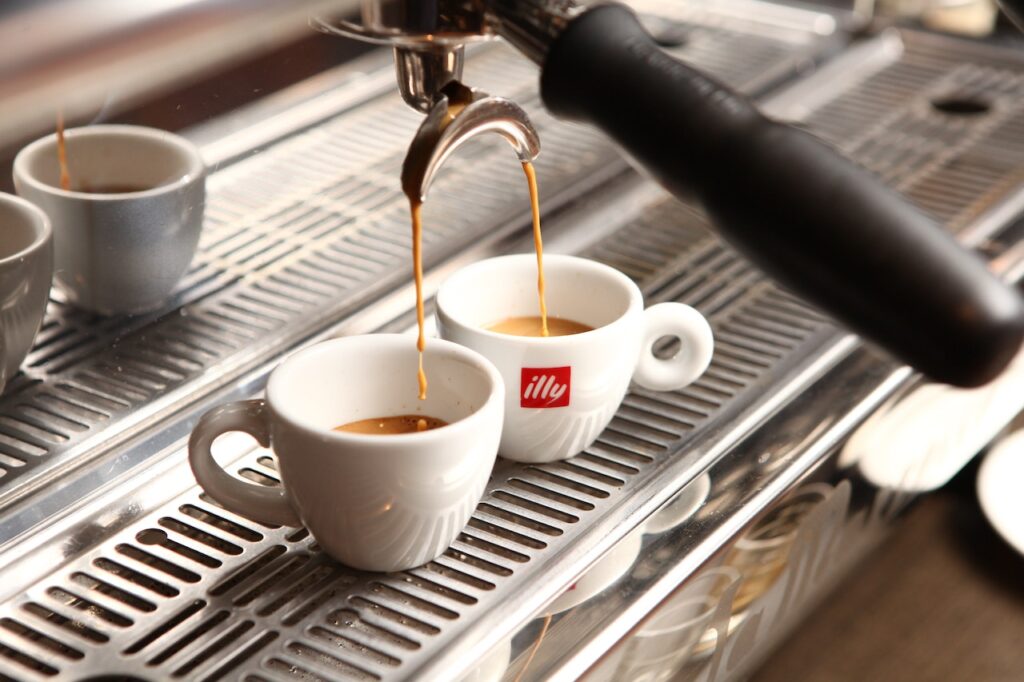 Two white Illy teacups are being filled with fresh coffee by a silver coffee machine in a coffee shop