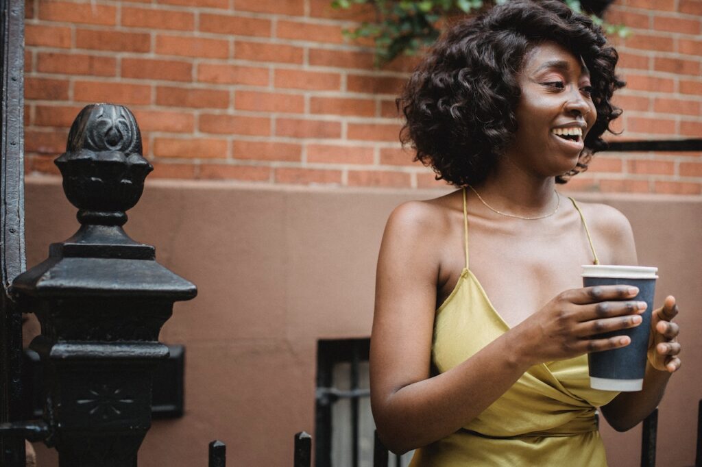 Is Coffee Bad for Your Teeth? 4 A woman with short curly hair wearing a yellow spaghetti strap dress holds a large cup of coffee outside the apartment