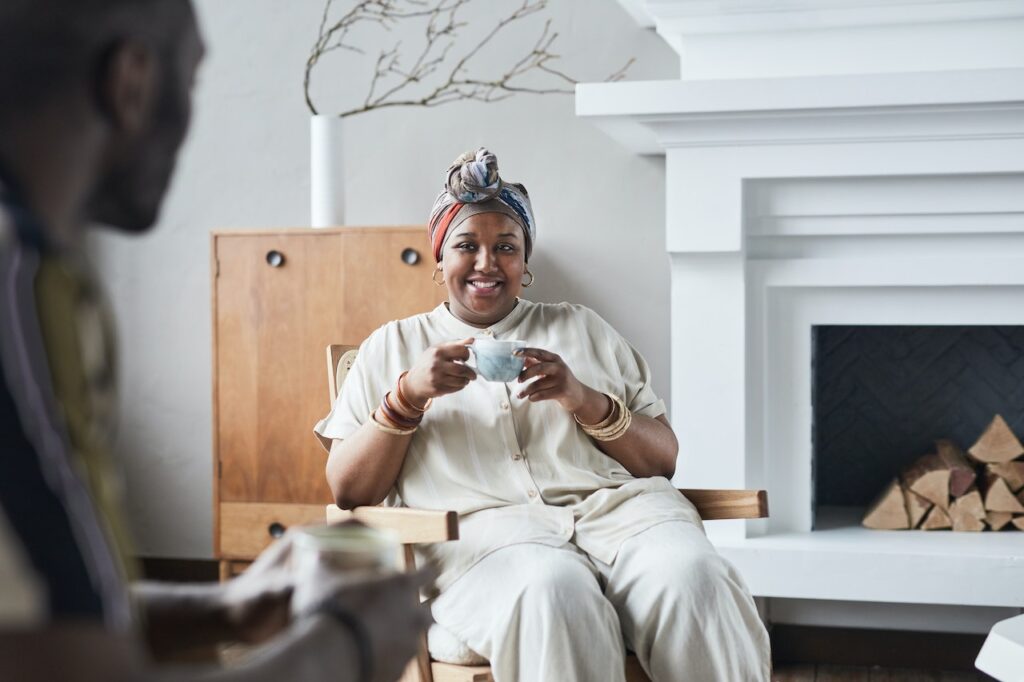 A woman wearing a colorful headscarf sits on a brown wooden chair near a white fireplace while holding a ceramic mug in the living room